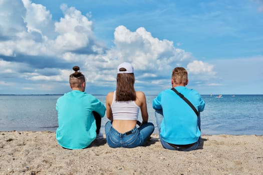 Back view of three teenage friends sitting on the sand at the beach, talking looking at sea. Youth guys and girl, summer vacation leisure friendship fun summertime happiness joy lifestyle holiday