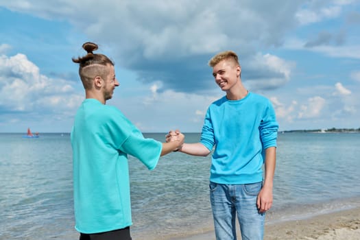 Young guys friends greet each other when they meet, shake hands, greeting, joy of meeting, outdoor on beach. Youth, communication, leisure, friendship, lifestyle concept