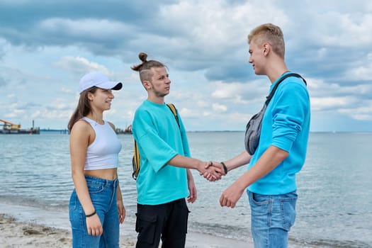 Young teenage friends guys and girl greet each other, shake hands, greeting, joy of meeting, outdoor on beach. Youth adolescence summer vacation communication leisure friendship fun lifestyle holiday