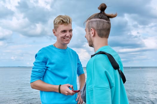 Two friends laughing cheerful young guys with smartphone, outdoor sea sky beach background. Men 18-20 years old age, youth, positive emotions leisure friendship communication, summer holidays concept