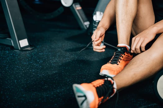 A fit and muscular male is tying his shoelace on the gym floor, surrounded by dumbbells and other workout equipment. The shot emphasizes his commitment to fitness and bodybuilding.