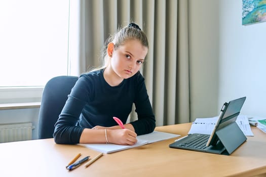 Portrait of smiling child girl sitting at desk at home with digital tablet textbooks, looking at camera. Education, knowledge, school, childhood concept