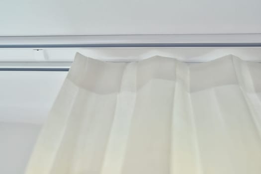 Translucent white textured curtain on window, close-up of ceiling cornice and fabric gathered on braid, laid and sewn with decorative folds, on plastic hooks