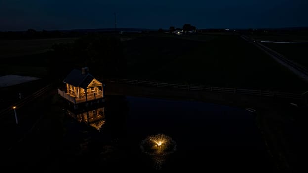 Ronks, Pennsylvania, USA, July 19 2024 - Tranquil Farm Scene At Dusk Featuring A Cozy Stone House Illuminated By Soft Lighting, Reflecting In A Peaceful Pond With A Fountain, Surrounded By Expansive Green Fields