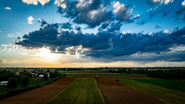 A vibrant sunset casts warm colors over expansive farmland, with lush green fields and patches of clouds creating a picturesque sky. The serene landscape reflects the tranquility of rural life.