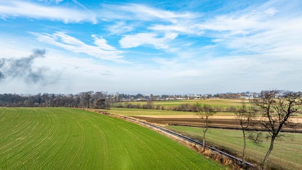 Expansive green fields stretch under a bright blue sky, adorned with a few clouds. Nearby, forests and distant farmlands showcase autumn hues, creating a serene rural landscape.