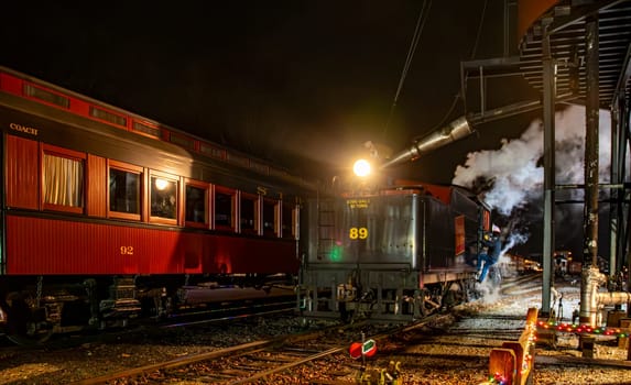 Strasburg, Pennsylvania, USA December 7, 2024 - Vintage steam locomotive is illuminated at night as engineers takes water on the locomotive. The surrounding area features a passenger car.