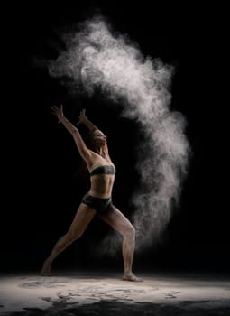 Full body fit woman in underwear making circle of sand in air while dancing against black background