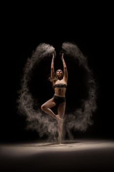 Full length slim female in bra and panties throwing sand in air and dancing against black background