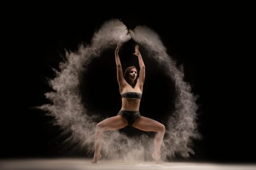 Full body fit woman in underwear making circle of sand in air while dancing against black background