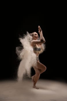Full length barefoot woman in underwear twisting arms and dancing in cloud of sand against black background