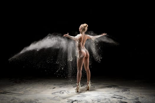 Back view seductive female in high heeled shoes throwing dust with outstretched arms against black background