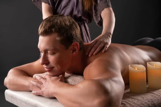 Focused professional masseuse doing massage on back of delighted male customer resting on table with candles in spa salon