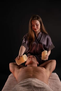 Professional masseuse applying melted wax from candles on chest of relaxed male client during spa procedure in salon