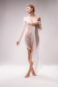 Seductive curvy female covering body with transparent cloth standing on tiptoes in studio on white background