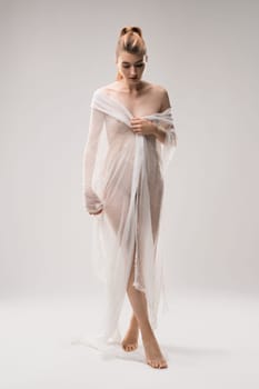 Graceful nude female covering body with transparent cloth standing on white background in studio and looking down