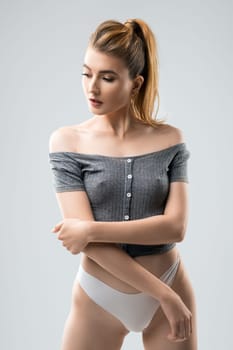 Sensual female model in stylish crop top and panties touching arm and looking away against gray background