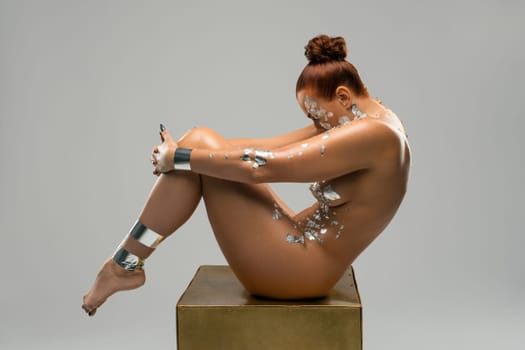 Full body side view of beautiful nude redhead female model with silver painting on perfect body posing on gold cube in light studio