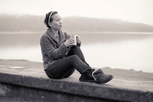 An Asian woman relaxes and enjoys a cup of coffee by the lake.