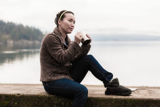 An Asian woman relaxes and enjoys a cup of coffee by the lake.