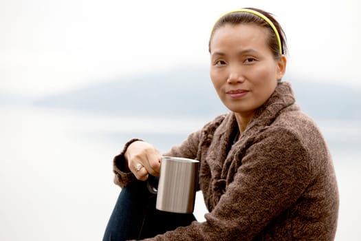 An Asian woman relaxes and enjoys a cup of coffee by the lake.