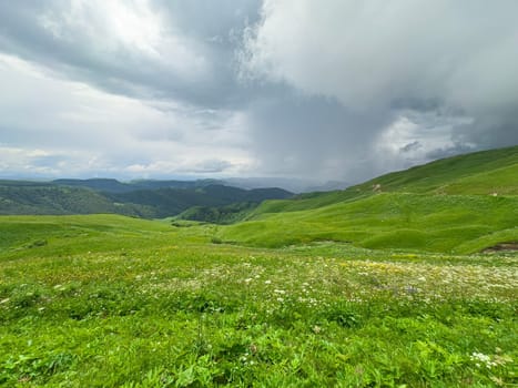 A vibrant green hillside stretches across the landscape, dotted with wildflowers. Dark storm clouds loom overhead, hinting at rain in this serene mountainous area.