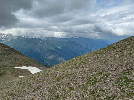 A vast mountain view with rocky terrain and patches of snow. Dark clouds loom overhead, framing the peaks and valleys of a remote alpine region, suggesting a change in weather.