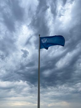 A vibrant blue flag flutters atop a tall pole under a swirling gray sky filled with clouds. The contrast between the flag and the turbulent weather creates a striking visual.