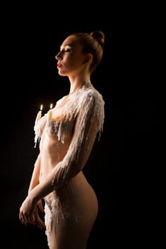 Nude female model with wax on shoulders closing eyes and burning candles on black background