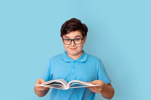 A cute schoolboy in glasses and a blue polo-shirt, holding an open book in his hands, look at camera, standing on a blue background. Copy space