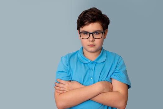 A saddark-haired boy with glasses, standing in a blue T-shirt, arms crossed on his chest, on a gray background, look at camera