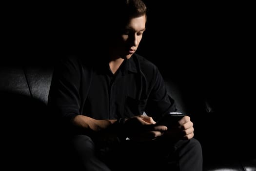 Concentrated young man in black outfit sitting on leather couch and browsing mobile phone in dark studio