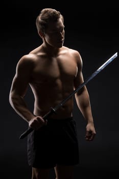 muscular male with naked torso holding Japanese katana sword in outstretched arm against black background