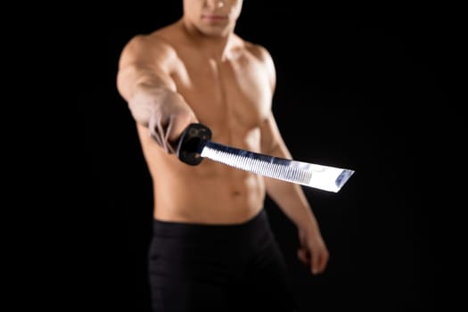 Crop unrecognizable muscular male with naked torso holding Japanese katana sword in outstretched arm against black background