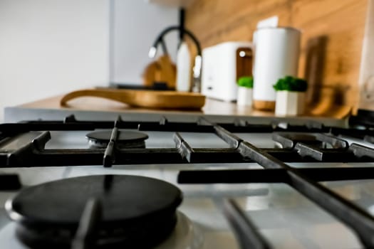 Close-up of a white gas stove with a black cast-iron grate and black burners against the background of a white and wooden kitchen out of focus. The concept of cooking, modern cuisine. High quality photo