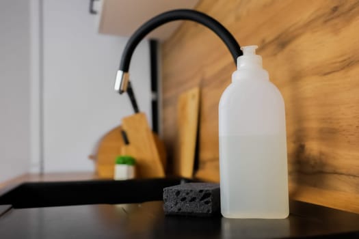 Close-up of a white bottle of dishwashing soap, a black dishwashing sponge lying next to it against the background of a black granite sink and a flexible high faucet and a white wooden modern kitchen out of focus. The concept of washing dishes, detergents, cleanliness. High quality photo