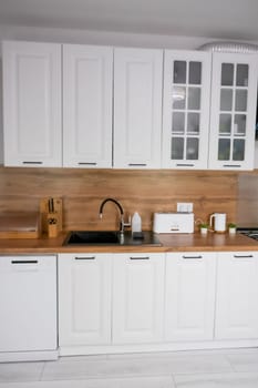 Horizontal photo of a modern kitchen in white and wooden style, in the center is a black kitchen sink, on a wooden countertop there are white wooden appliances for storing and preparing food. Modern kitchen concept, kitchen island, modern kitchen style. High quality photo