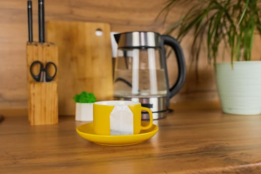 A yellow cup of tea stands on a yellow saucer and on the saucer lies a tea bag on a wooden table against the background of a teapot, a plant and kitchen utensils. The concept of tea drinking, break. High quality photo