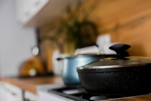 Close-up of a black frying pan with a transparent lid and a blue saucepan with a transparent lid and silicone pads on the handles against the background of a modern wooden-white kitchen out of focus. The concept of cooking, cooking, boiling and frying. High quality photo