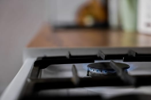 Close-up of a white gas stove with a black cast-iron grate and a black burner on which a blue fire burns against the background of a wooden kitchen in out-of-focus. Gas stove, cooking concept. High quality photo