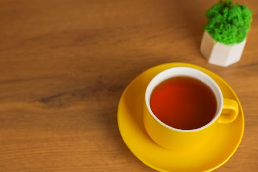 A top view of a yellow cup with a saucer in which black tea is poured stands on a wooden table on the left with space for text, next to it there is a plant in a white pot. Concept of tea drinking, cleaning break, rest. High quality photo