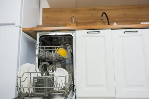 An open dishwasher with a dish drawer pulled out close-up on the left, it contains clean dishes and cutlery against the background of a white wooden kitchen. Comfort concept, dishwashing. High quality photo