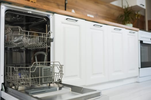 An empty open dishwasher with gray drawers for dishes and cutlery stands on the left of the photo against the background of a white wooden kitchen in a slight out of focus. Dishwashing concept, comfort. High quality photo