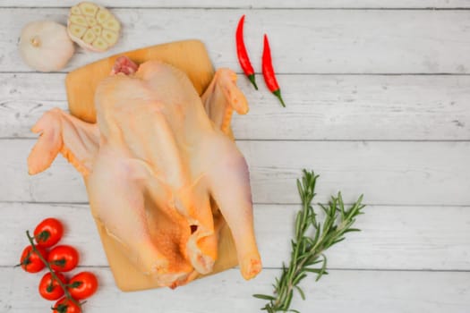 Close-up top view of a butchered raw chicken lying on a wooden cutting board surrounded by branches of red tomatoes, garlic, red hot peppers, sprigs of rosemary against a white tree background with space for text. The concept of cooking, diet menu, ingredients. High quality photo