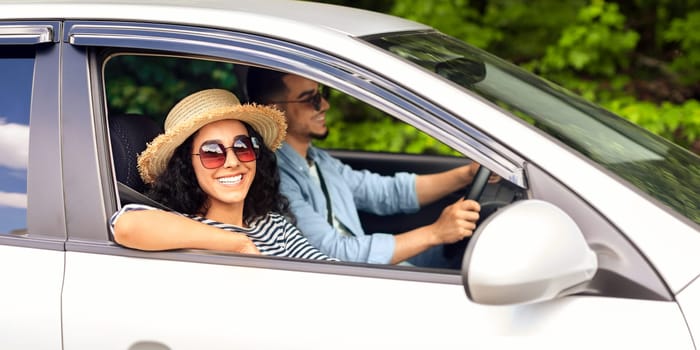 Smiling young lady in summer hat and sunglasses looking at camera while having car trip with her man, middle-eastern couple going vacation together, enjoying nice weather, side view
