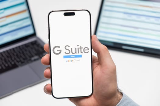 PRAGUE, CZECHIA - JULY, 2025: G Suite logo on the screen of smartphone in mans hand and laptop on the background