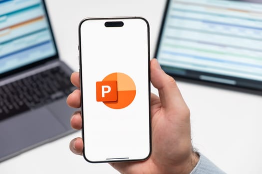 PRAGUE, CZECHIA - JULY, 2024: Power Point logo on the screen of smartphone in mans hand and laptop on the background
