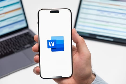 PRAGUE, CZECHIA - JULY, 2024: Word logo on the screen of smartphone in mans hand and laptop on the background