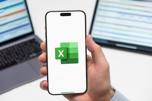PRAGUE, CZECHIA - JULY, 2024: Excel logo on the screen of smartphone in mans hand and laptop on the background
