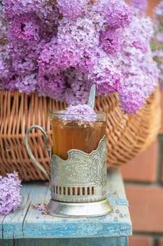 Glass of tea with lilac flowers in a traditional russian teacup holder standing on a wooden surface next to a wicker basket full of lilac flowers, creating a cozy spring atmosphere